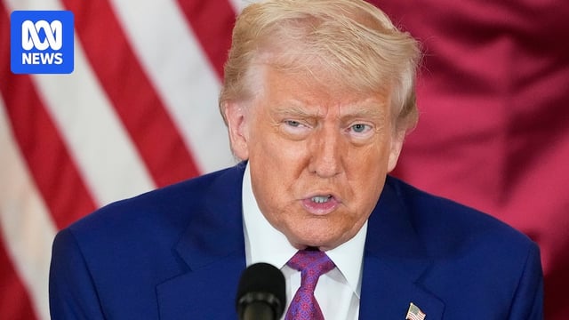Donald Trump wearing a blue suit and purple tie with a US flag lapel pin, speaking in front of a US and Qatari flag