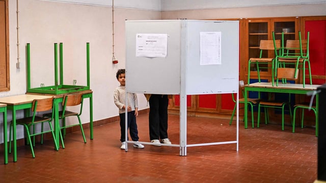 Ein Kind schaut hinter einer Wahlkabine hervor.