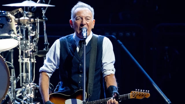 A white man stands at a microphone while playing a guitar. Part of a drumset can be seen behind him.