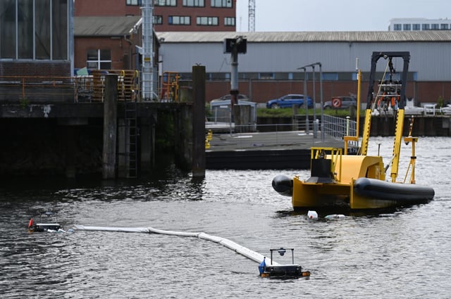 Zwei schwimmende Roboter sammeln bei einem Pressetermin in Hamburg-Harburg Müll auf dem Wasser ein, im Hintergrund fährt ein Schiff, dass den Müll aufsammeln soll.