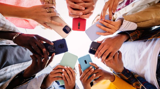 A group of teenagers stand in a tight circle, each holding up a smartphone with colorful cases. The shot is taken from below, capturing the sense of collective immersion in their screens. The image reflects the growing entanglement of youth with digital platforms and AI-driven technologies, echoing concerns raised in the Florida lawsuit involving a chatbot and a 14-year-old's tragic suicide.