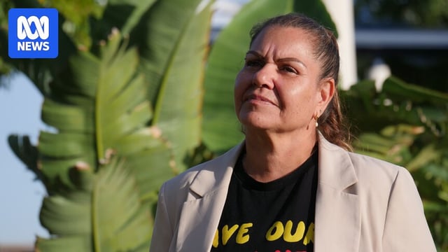 An australian aboriginal woman wearing a beige jacket and black shirt stares into the sky, with banana leaves behind her
