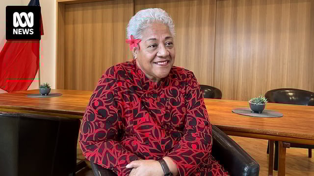 A Samoan woman sitting in red and black patterned outfit with short white hair and red flower behind her ear.