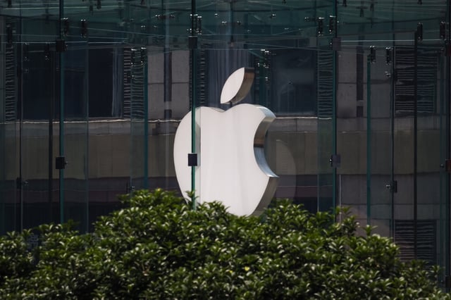 The Apple logo is displayed on the glass facade of an Apple Store, partially obscured by green foliage in the foreground, on May 20, 2025 in Chongqing, China.