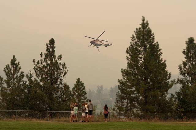 People gather to a lookout as a helicopter gathers water from Shannon Lake to fight the McDougall Creek wildfires in West Kelowna, British Columbia on August 19, 2023.