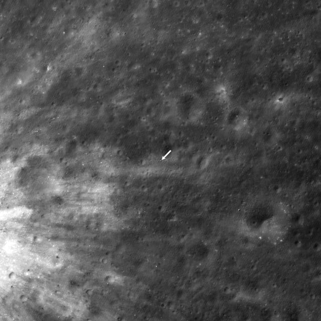 A black and white image of the surface of the moon taken from the Lunar Reconnaissance Orbiter. A white arrow in the center of the image points to the location of the SLIM lander.