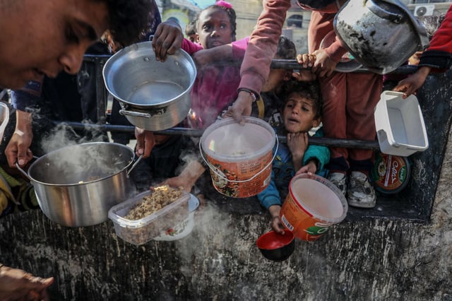 Palestinian children wait in line to receive food prepared by volunteers for Palestinian families displaced to Southern Gaza due to Israeli attacks, between rubble of destroyed buildings in Rafah, Gaza, on February 9, 2024.