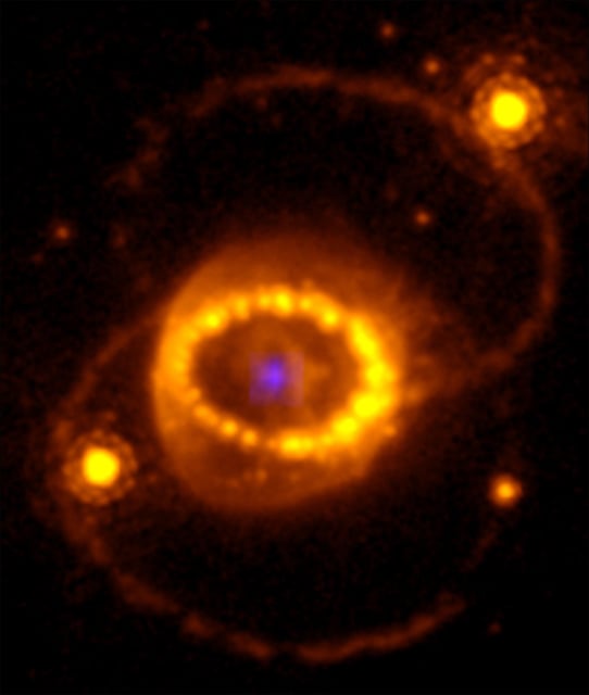Figure 1. Combination of a Hubble image of SN 1987A and the compact highly ionized argon source in Fig. 2. The faint blue source in the centre was detected by the NIRSpec instrument on JWST. Outside of this is the rest of the supernova, which contains the most mass and is expanding at thousands of kilometers every second. The inner bright "string of pearls" is gas from the star's outer layers that was ejected about 20,000 years before the explosion. The collision between the rapidly expanding supernova remnant and the ring gives rise to the heated clumps in the ring. Outside the inner ring are two outer rings, which probably arose at the same time as the inner ring was formed. The bright stars to the left and right of the inner ring are unrelated to the supernova. (Illustration: HST, JWST/NIRSpec, J. Larsson)