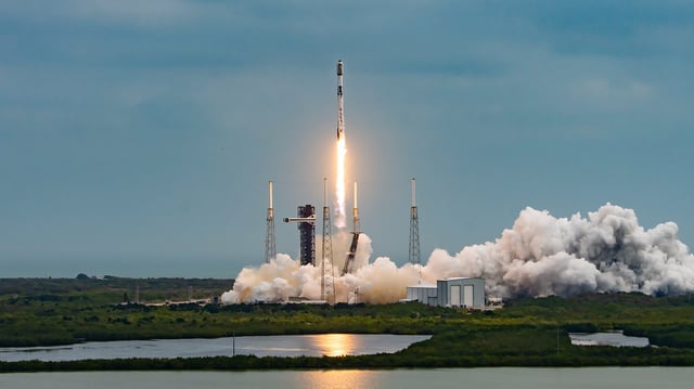 a white rocket launches above lush green wetlands during the day