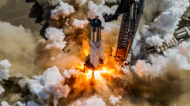flames billow from the base of a large silver rocket that's sitting on a test stand.