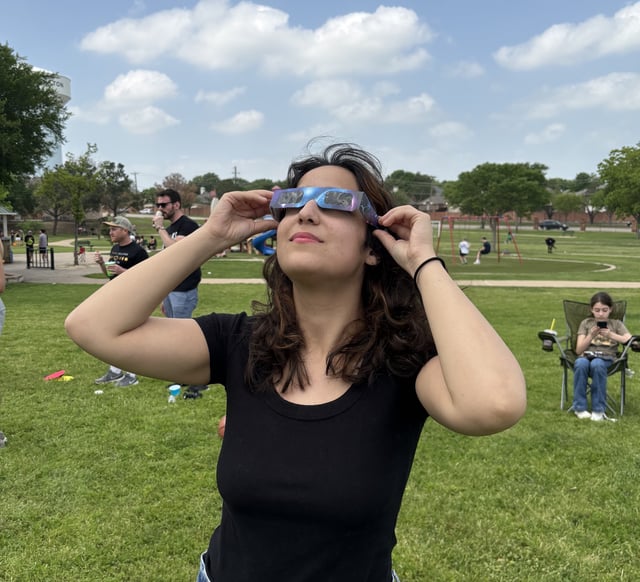 Emma Friedman, a NASA intern, is pictured here observing the total solar eclipse on April 8, 2024. Emma is standing on a grassy field wearing a black shirt and special eclipse sunglasses. A blue sky can be seen behind her.