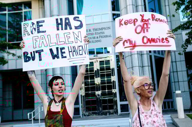 Two women hold signs reading "ROE HAS FALLEN BUT WE HAVEN'T" and "KEEP YOUR ROSARIES OFF MY OVARIES" during an outdoor demonstration