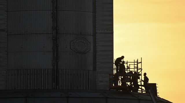 workers on scaffolding in hard hats next to a giant metal rocket