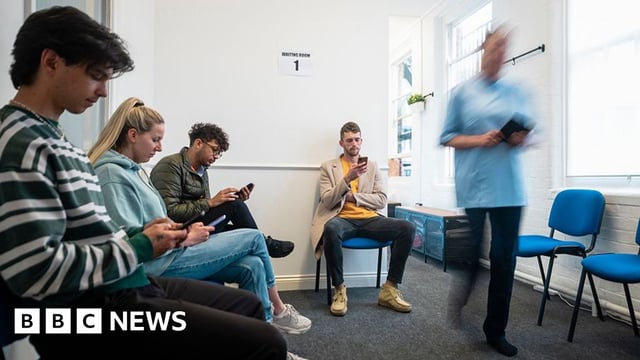 People wait in a healthcare waiting room as a healthcare professional walks though