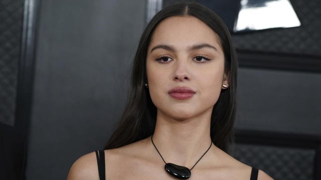 Olivia Rodrigo posing in a black, chunky necklace against a black background