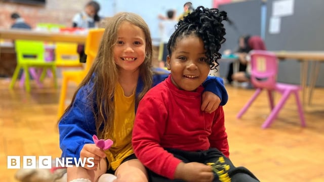 A child with long, light brown hair, and a yellow and blue top, has her arm around another child with black curly hair. They're both smiling and sitting on a wooden floor inside a holiday club.
