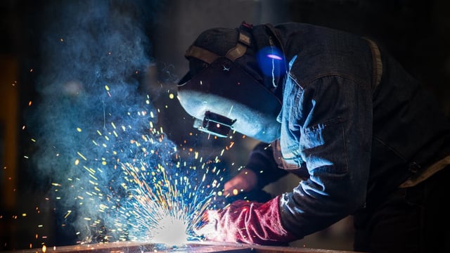A photograph of a person welding. They are wearing a mask and protective clothing and gloves. There are sparks and fumes being emitted from the weld.