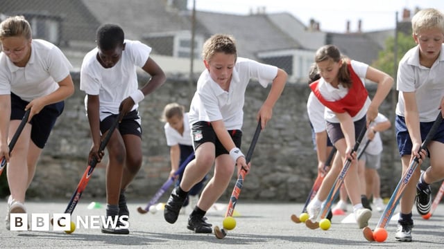 A row of pupils in white and black sports PE kits carefully guiding small balls with hockey sticks with a grey stone wall in the background