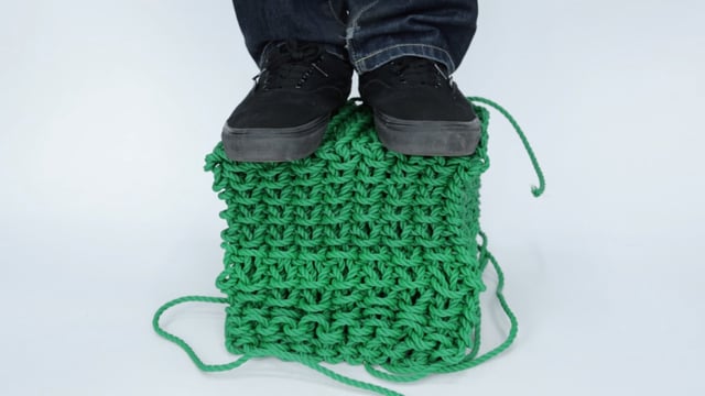 A green cube made out of knitted cord is being stood on by a person wearing dark jeans and black sneakers