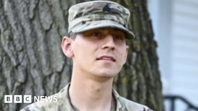 Soldier in a military cap and fatigues stands in front of a tree.