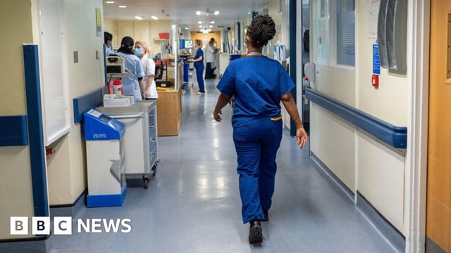 A doctor wearing dark blue scrubs and a face mask, with her back to the camera, walks down a hospital corridor