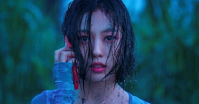 Ko Min-si holding up a red mobile phone to her ear while standing in the middle of a forest in the rain.
