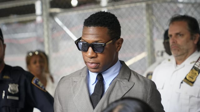 Jonathan Majors is leaving a courthouse while wearing a gray suit with a black tie and sunglasses