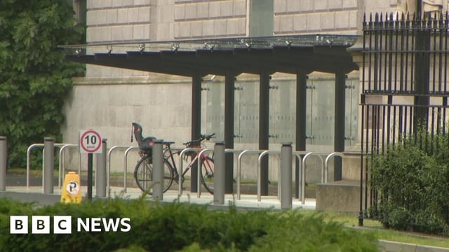 A metal and glass bike shelter with metals railings to prop bicycles against. One bike is in the shelter. Behind the shelter a grey building