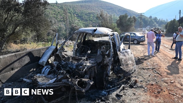 A damaged burnt out car on the outskirts of Masyaf lies on the side of a dirty road in Syria. People watch on and take photos of the damaged car.