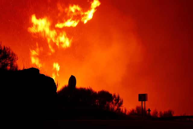 A cross on the road is silhouetted, as flames and smoke coming from a wildfire rise near Povoa de Montemuro, Portugal, September 18, 2024. REUTERS/Susana Vera