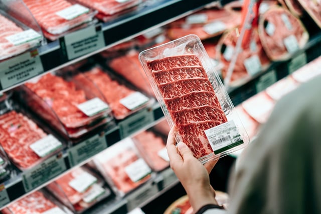 Over the shoulder view of young Asian woman shopping in a supermarket. She is choosing meat and holding a packet of organic beef in front of the refrigerated section.